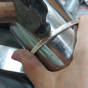 May include: A person is using a hammer to shape a silver bracelet on a metal form. The bracelet is being hammered into a curved shape.