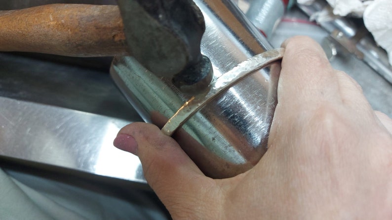May include: A person is using a hammer to shape a silver bracelet on a metal form. The bracelet is being hammered into a curved shape.