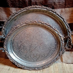 Op de afbeelding: Twee sierlijke zilverkleurige trays met ingewikkelde bloempatronen. De trays zijn ovaal en rond, met decoratieve handvatten en randen. De trays zijn in elkaar genest. De achtergrond is een gestreepte stof.