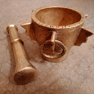 Peut inclure: Un mortier et un pilon en or avec des motifs complexes. Le mortier a une base large et ronde et un bol profond. Le pilon est de forme cylindrique longue avec une extrémité arrondie.