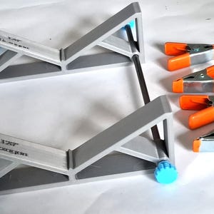 May include: A grey, angled woodworking jig with blue adjustment knobs and black support rods. The jig is labelled "120° Hexagon." Five orange and silver spring clamps are positioned to the right, providing a complete set for woodworking projects.