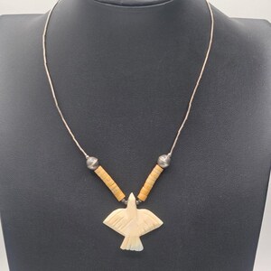 May include: A necklace featuring a carved off-white bird pendant suspended from a thin silver chain. The design includes two sections of cylindrical tan beads and two spherical silver beads positioned above the bird-shaped focal point.