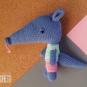 May include: A blue crocheted anteater toy wearing a green and pink outfit. The anteater has a long snout and a pink tongue sticking out.