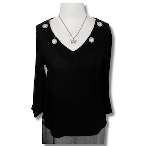 May include: Black long-sleeved blouse with a V-neckline and silver button accents. The blouse has a butterfly pendant necklace.