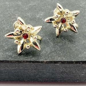 Peut inclure: Une paire de boucles d'oreilles à clous en métal doré en forme d'étoiles à cinq branches ou de fleurs. Chaque boucle d'oreille présente une petite pierre précieuse rouge circulaire au centre, entourée de pétales métalliques et de perles.