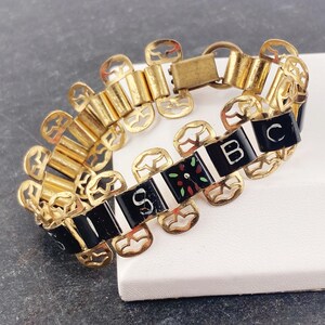 May include: A gold-toned bracelet with black and white rectangular links. The bracelet features the letters "SBC" and decorative floral accents in red, green, and black. The bracelet has a clasp closure.