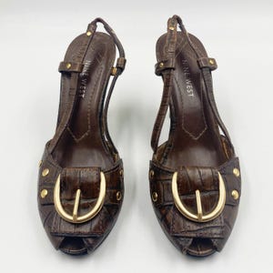 May include: A pair of brown Nine West slingback heels featuring a faux crocodile skin texture, an open toe, and a prominent gold-toned buckle detail on the front. The shoes include gold-toned studs on the sides and a thin adjustable heel strap.