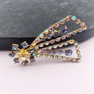 May include: A gold-tone metal brooch shaped like a shooting star, featuring a central cluster of round blue and iridescent rhinestones. The tail of the star consists of multiple curved lines set with shimmering blue and clear crystal stones.