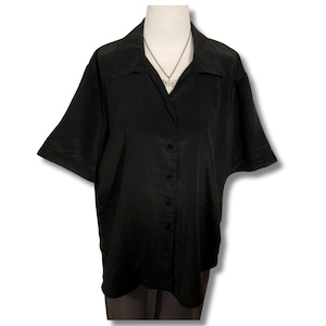 May include: A black button-down shirt with short sleeves and a collar. The shirt is displayed on a mannequin, with a silver necklace visible. The fabric appears to be a smooth, possibly silk or satin, material.