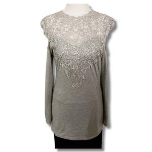 May include: A gray long-sleeved shirt with a detailed, light-colored lace overlay on the upper chest and shoulders. The lace design is intricate and adds a decorative element to the top. The shirt has a simple, classic design.