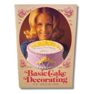 Op de afbeelding: Vintage boekomslag getiteld "Basic Cake Decorating" met een vrouw die een versierde taart vasthoudt. De taart is roze met witte glazuur en gele rozen. De omslag heeft een bloemenrand en de tekst "THE WILTON WAY".