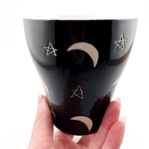 May include: A black ceramic mug with a white interior. The exterior features etched crescent moon and star designs. The mug is held in a hand against a white background. The mug has a tapered shape.