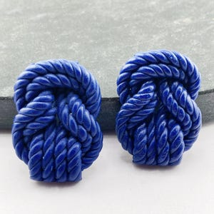Op de afbeelding: Twee marineblauwe oorstekers in de vorm van een touwknoop. Elke oorbel heeft een gedetailleerde touwtextuur en een glanzende afwerking. Ze worden gepresenteerd tegen een neutrale achtergrond, waardoor hun levendige kleur en nautische thema worden benadrukt.