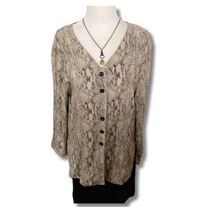 May include: A button-down blouse with a snakeskin pattern in shades of beige and gray. The long-sleeved top has a V-neck and black buttons. A black necklace with a gold pendant is also visible.