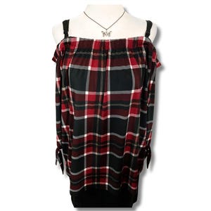 May include: Off-the-shoulder top in a red, black, and white plaid design. The long-sleeved top features black straps and wrist ties. A silver butterfly pendant hangs from a delicate chain. The top is displayed on a mannequin.