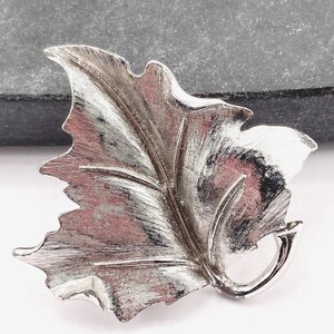 May include: Silver-coloured leaf-shaped brooch with detailed veining. The brooch has a textured surface and a curved stem. The design is reminiscent of a maple leaf. A classic accessory for adding a touch of nature-inspired elegance.