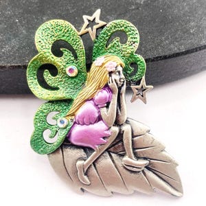 May include: A detailed fairy brooch featuring a seated fairy with blonde hair and a pink dress, resting on a silver leaf. The fairy has green and yellow wings with star accents and iridescent gems. A whimsical accessory.