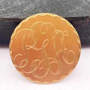 Pode incluir: Um disco metálico circular de tom dourado com um monograma intrincado gravado à mão com as letras R, S e H num estilo de escrita fluido. A borda do disco é finalizada com um padrão decorativo recortado, criando uma borda texturizada.