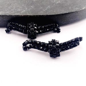 May include: Two black rhinestone embellished bow-shaped jewelry components. The bows are made of small, square-cut black rhinestones, giving them a sparkling appearance. The bows are displayed on a white surface with a dark gray background.