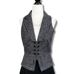 May include: A sleeveless charcoal gray tweed vest featuring a notched lapel collar, a double-breasted front closure with four black buttons, and two rectangular flap pockets at the waist. The garment is displayed on a white mannequin torso.