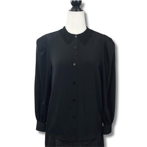 May include: A black long-sleeved button-down shirt featuring a delicate black lace collar. The garment has slightly puffed shoulders, long sleeves with buttoned cuffs, and a central vertical row of matching black buttons down the front.