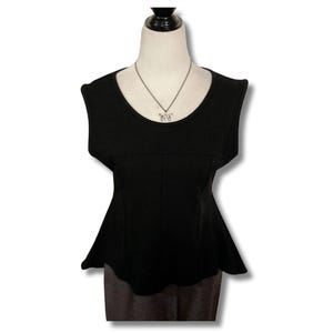 May include: A black sleeveless top with a peplum hemline, displayed on a mannequin. The top has a scoop neckline and a fitted bodice. A silver necklace with a small pendant is visible. The mannequin is wearing a gray skirt.