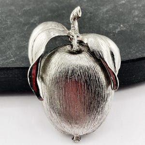 Puede incluir: Un broche de tono plateado con forma de manzana y acabado de metal cepillado texturizado. La pieza presenta un cuerpo central redondeado coronado por un tallo corto y dos hojas curvas prominentes que se extienden a ambos lados.