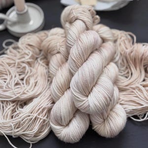May include: Close-up of beige yarn skeins and loose yarn. The yarn has a soft, textured appearance with subtle variations in color. The skeins are tightly wound, while the loose yarn is spread out, suggesting a crafting project.