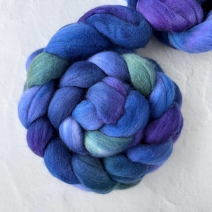 May include: Close-up of a braid of colorful wool roving in shades of blue, teal, and purple. The wool is soft and fluffy, perfect for fiber art projects like spinning, felting, or weaving. The roving is arranged on a white surface.