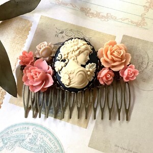 May include: A gold hair comb with a black cameo featuring a woman's profile and a cluster of pink and peach fabric flowers.