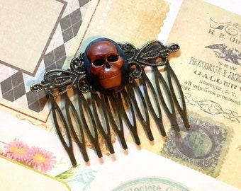 May include: A brown skull hair comb with a decorative metal design. The comb has a dark brown skull with a white face and black eyes. The comb is on a background of vintage paper with text.