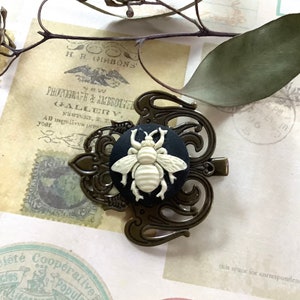 May include: A black and white cameo featuring a bee with intricate details, set in a bronze filigree frame. The cameo is attached to a hair clip.