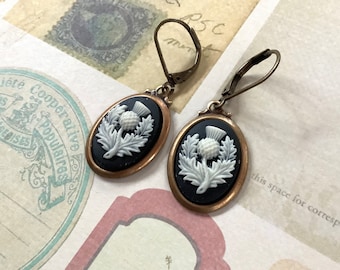 May include: A pair of earrings with a black and white cameo featuring a stylized thistle design. The earrings are made of bronze and have a hook closure.