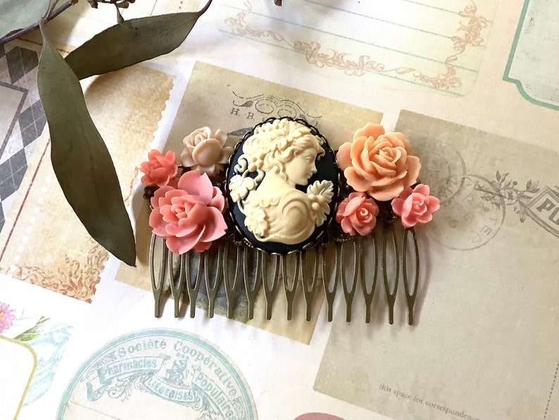 May include: A gold hair comb with a black cameo featuring a woman's profile and a cluster of pink and peach fabric flowers.
