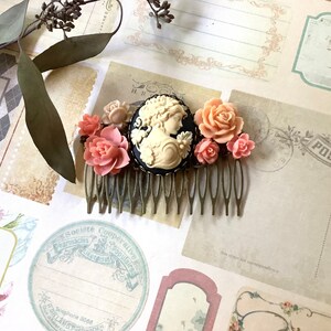 May include: A gold hair comb with a cameo of a woman's profile in a black frame. The comb is decorated with pink and white flowers.