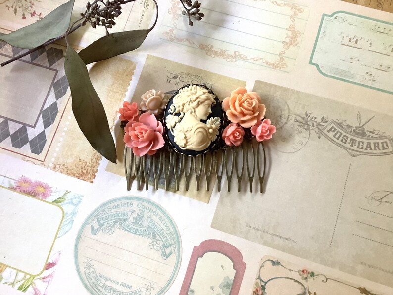 May include: A gold hair comb with a cameo of a woman's profile in a black frame. The comb is decorated with pink and white flowers.