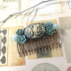 May include: A silver hair comb with a cameo featuring three mermaids in a seashell. The cameo is surrounded by blue and white flower accents.