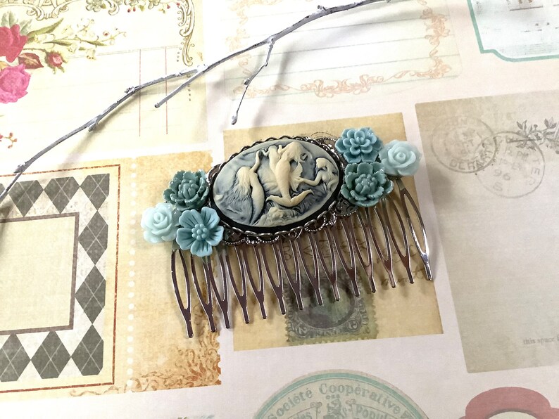 May include: A silver hair comb with a cameo featuring three mermaids in a seashell. The cameo is surrounded by blue and white flower accents.