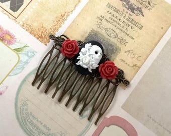 May include: A bronze hair comb with a black skull cameo and three red flower accents. The skull cameo has white floral details. The comb has seven teeth.