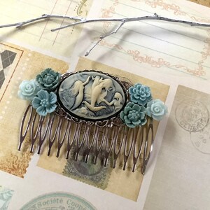 May include: A silver hair comb with a cameo featuring a white porcelain design of three mermaids. The comb is decorated with blue and green flower accents.