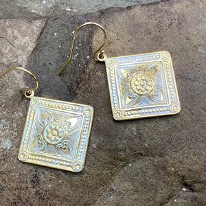 May include: A pair of gold-toned square dangle earrings with an intricate floral design. The earrings have a textured surface and a delicate hook closure.