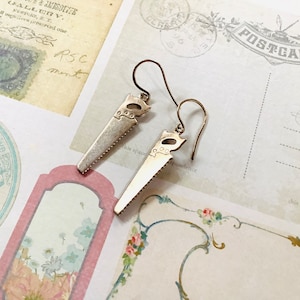 May include: A pair of silver earrings shaped like a saw. The earrings are hanging from a white background.