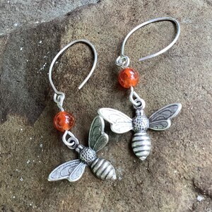 Könnte beinhalten: Silberne Bienenohrringe mit orangefarbenen Karneolperlen. Die Bienen haben detaillierte Flügel und Körper.