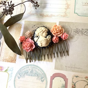 May include: A gold hair comb with a black cameo featuring a woman's profile. The comb is decorated with pink and white plastic flowers.