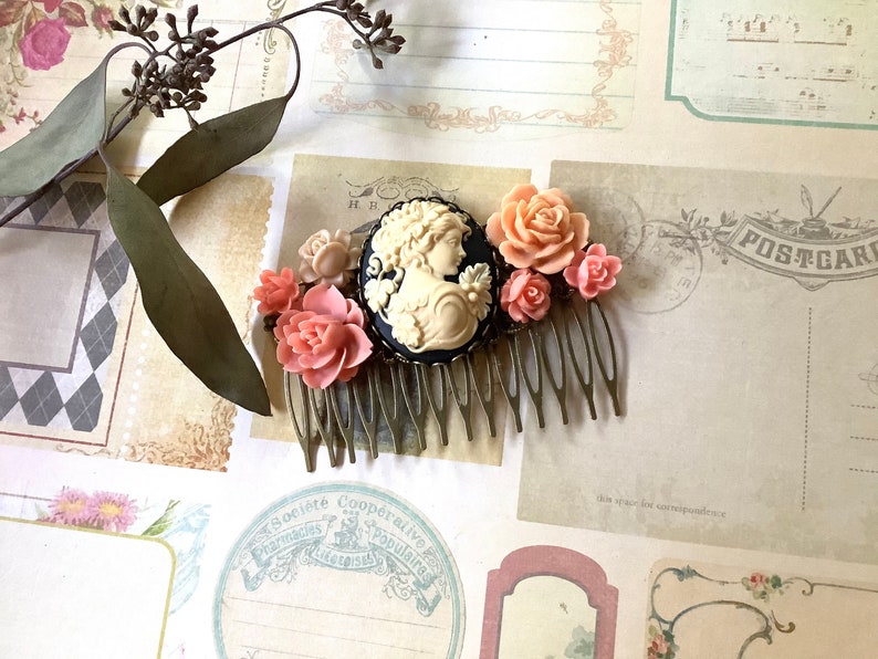 May include: A gold hair comb with a black cameo featuring a woman's profile. The comb is decorated with pink and white plastic flowers.