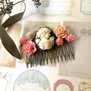 May include: A gold hair comb with a black cameo featuring a woman's profile and a cluster of pink and cream colored flowers.