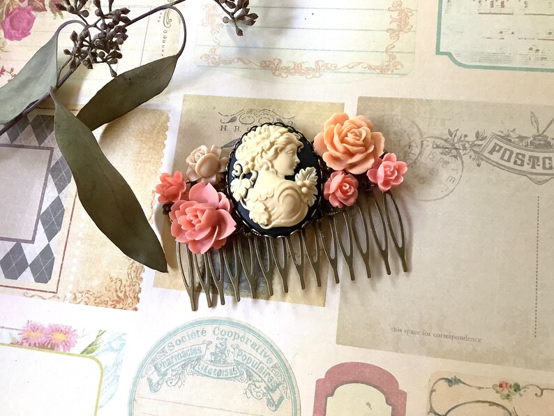 May include: A gold hair comb with a black cameo featuring a woman's profile and a cluster of pink and cream colored flowers.