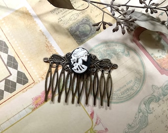 May include: A bronze hair comb with a black and white cameo featuring a woman's profile. The comb has a decorative design with swirls and loops.