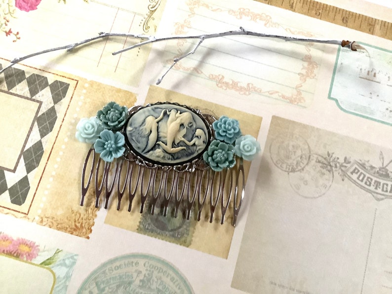 May include: A silver hair comb with a cameo featuring a white and blue mermaid design. The comb is decorated with light blue and white flowers.