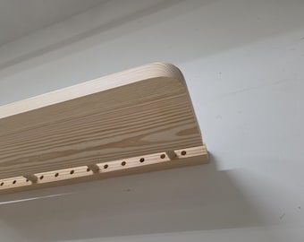 May include: A light-colored wooden wall shelf featuring a smooth, rounded corner edge and a horizontal wooden strip attached to the front. This strip contains a series of evenly spaced circular holes and small wooden pegs extending outward.